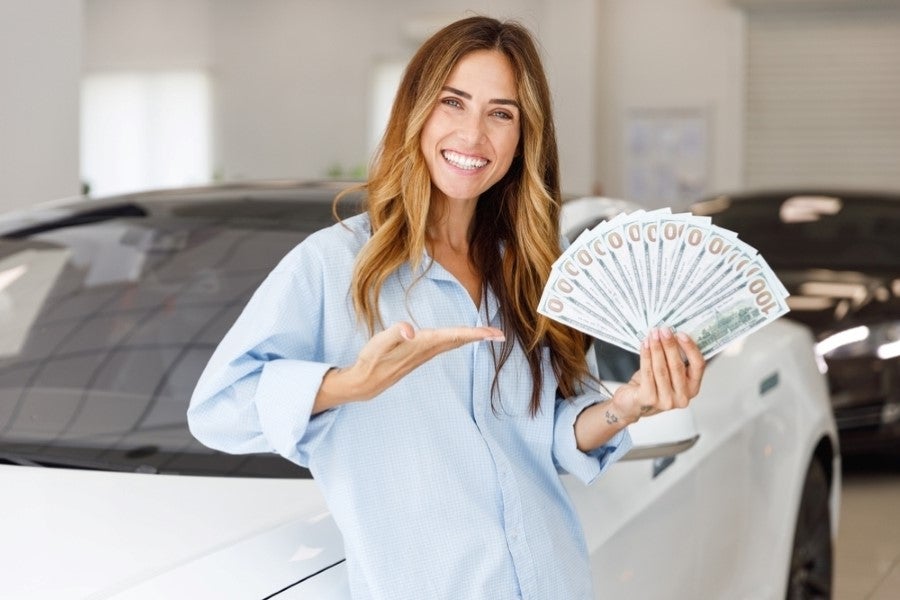 A smiling woman stands in front of a car, holding and pointing to a fan of cash.